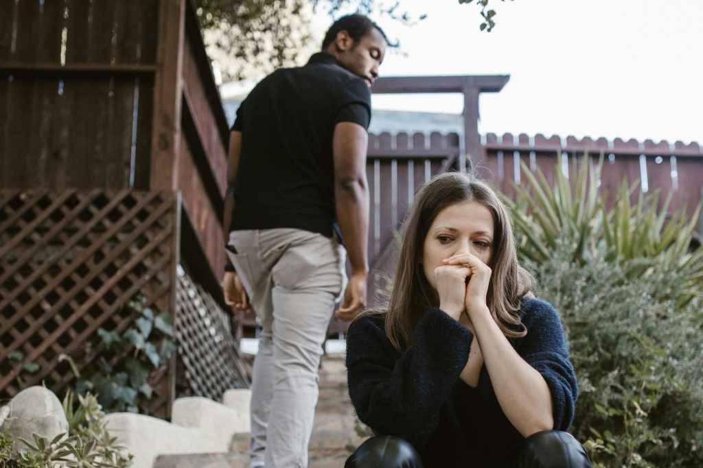 Woman sitting on steps looking worried with hand on her face, while man walks away up the steps and looks back at her